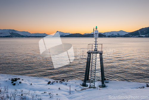 Image of Snow-covered coastline with a navigation beacon overlooking a tranquil sea during a scenic sunset.