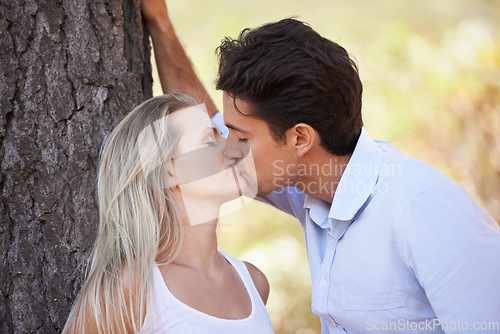 Image of Couple, kiss and love by tree for affection, care or romance in nature, outdoor park or forest. Young man and woman kissing for embrace, relationship or bonding intimacy in woods for date or freedom