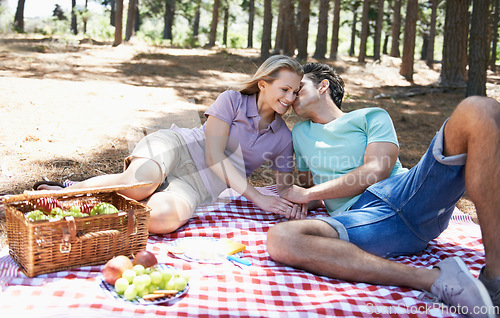 Image of Happy couple, kiss and relax in nature for picnic, love or support in affection, date or outdoor bonding. Woman and man sitting on floor with basket of fruit for embrace, eating or romance in forest