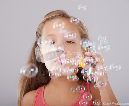 Image of Portrait, little girl and blowing bubbles in studio by toy, cute and fun games with soap liquid. Child, face and learning to play with bubble wand, childhood development and sweet by gray background