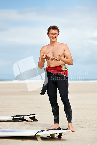 Image of Surfer, man and face or happy with surfboard on beach with wetsuit, blue sky and confidence with sunglasses. Extreme sports, athlete and person by ocean for training, surfing workout and smile