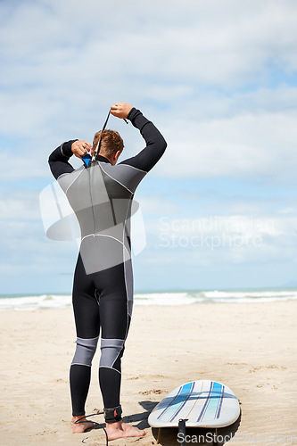 Image of Surfer, man and getting ready with surfboard on beach with wetsuit, blue sky and dressing with mock up space. Rear view, athlete and person by ocean for training, surfing workout and extreme sports