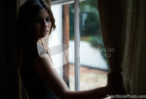 Image of Woman, portrait and window at night in dark room in stress, anxiety or mental health for lonely indoor home. Young female person standing by glass at house by curtains in neighborhood apartment