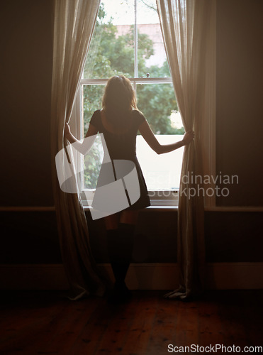 Image of Woman, curtain and morning by window for sunlight, vitamin D or sunshine in bedroom at home. Rear view of female person standing by blinds or glass in casual clothing and looking out in neighborhood