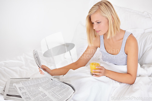 Image of Young woman, orange juice and reading newspaper on bed for headlines, energy and health body with wellness. Blonde person, thirsty and glass of fruit drink to detox, morning and vitamin c in bedroom