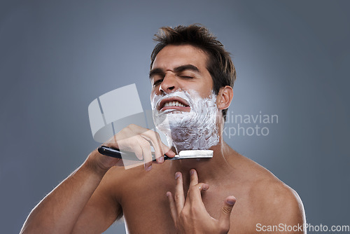 Image of Man, shaving and razor with stress in studio for grooming, skincare and wellness with pain by grey background. Person, model and annoyed with beard, facial hair removal and cream for cosmetic change