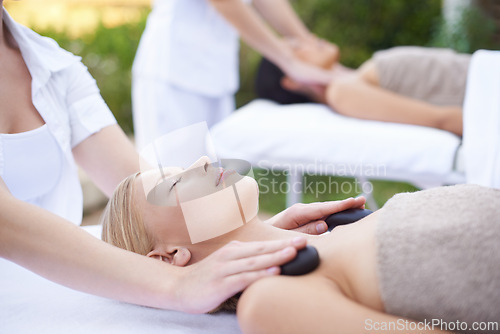 Image of Women, hands and massage with hot stone in spa, outdoor and wellness on bed for body treatment. People, heat and masseuse for release of muscle tension, nature and health detox in comfort at resort