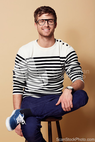 Image of Portrait, fashion and glasses with a man on a chair in studio on tan background for trendy style. Relax, casual and eyewear with a confident young geek or nerd sitting on a stool in a clothes outfit