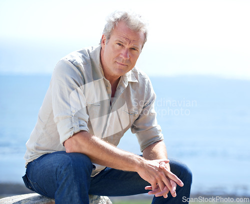 Image of Mature, man and outdoor portrait at the sea on holiday, vacation or travel in nature to relax. Serious, person and calm ocean, water or waves in morning and sitting on a rock with blue sky in summer