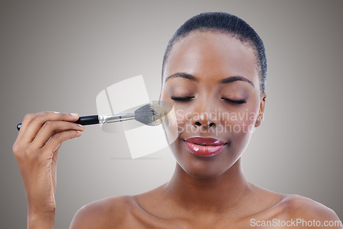 Image of Face, beauty and makeup brush with young black woman in studio on gray background to apply blush to cheek. Aesthetic, product and foundation with confident model using luxury cosmetics for makeover