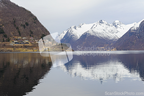 Image of A tranquil fjord with a village on the shore, reflecting snow-covered mountains in calm water.