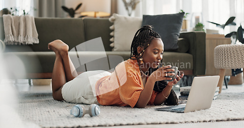 Image of Laptop, coffee and woman on the floor in living room of modern apartment reading information. Technology, relax and young African female student study and drink latte on computer in lounge at home.