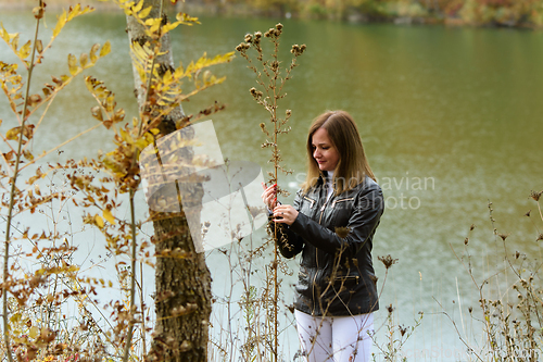 Image of A girl stands by the lake on a warm autumn day and examines the leaves of plants.