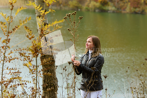 Image of A girl stands by the lake on a warm autumn day and holds a tall dry bush