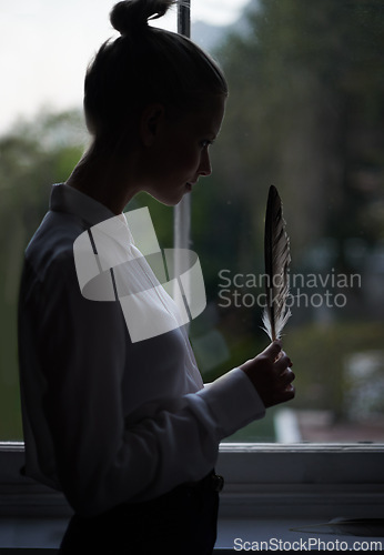 Image of Woman, feather and window for writer in home, dark room and creative for abstract or artistic pose. Female artist, writing and shadow with thinking, fashion and mysterious editor for literature