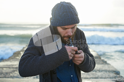 Image of Bearded, man and smoking a pipe by ocean, lighter and tobacco habit on winter morning for sunrise. English guy, nicotine and vintage smoker for calm, satisfaction and vacation on beach in cape town