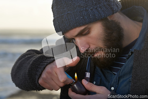 Image of Bearded, man and smoking a pipe on beach, lighter and tobacco habit on winter morning for sunrise. English guy, nicotine and vintage smoker for calm, satisfaction and vacation by ocean in cape town
