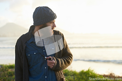Image of Man, pipe and thinking in nature by ocean for ideas, vision or planning on winter morning. Male person, sunrise and standing on grass by sea for relaxation, wondering or smoking with tobacco outdoors