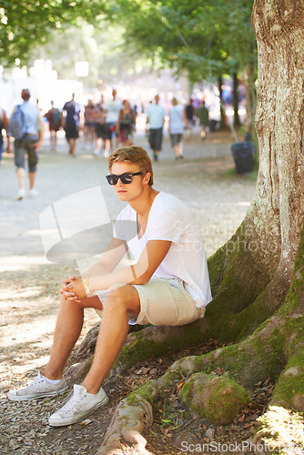 Image of Nature, sunglasses and man sitting by tree with confidence in an outdoor park on vacation. Travel, garden and male person thinking by wood trunk in field on holiday, adventure or weekend trip.