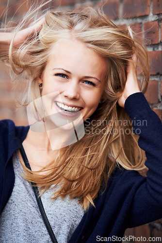 Image of Portrait, happy and woman with wind in hair by brick wall in city, funny or laugh outdoor. Face, fashion and smile of person, stylish casual clothes and blonde girl with breeze in Switzerland