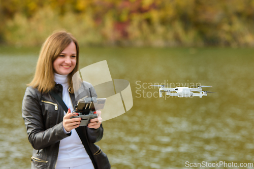 Image of A young beautiful girl launches a radio-controlled quadcopter on the shore of an autumn lake, the girl looks at the drone smiling
