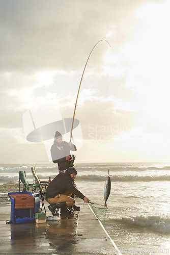 Image of Fishing, men and net with fish at pier with rod, waves and relax on vacation, holiday and adventure. Friendship, people and bonding in morning with overcast, sky and nature for activity and hobby