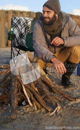 Image of Campfire, wood and man by beach on vacation, adventure or holiday camping for travel. Nature, outdoor and young male person sitting on chair in sand with flame for heat on weekend trip in winter.