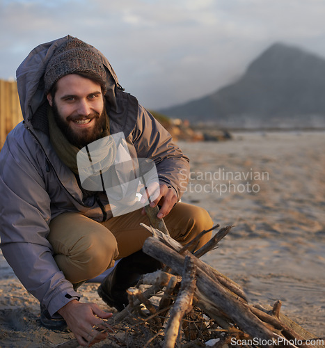 Image of Portrait, smile and man at beach with campfire on holiday, vacation or travel toe prepare for winter outdoor alone. Happy person, wood and adventure at seashore, leisure and recreation in Canada