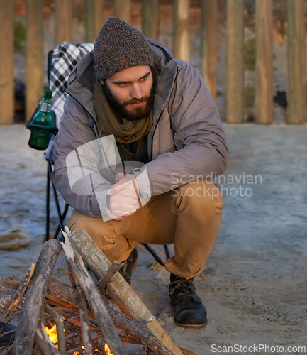 Image of Campfire, wood and man by beach for travel on vacation, adventure or holiday camping. Nature, outdoor and young male person sitting on chair in sand with flame for heat on weekend trip in winter.