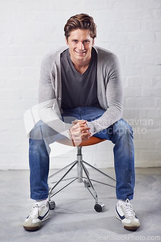 Image of Man, portrait and smile on chair with fashion on wall background for style, confidence or relax. Male person, face and sitting or casual in jeans in studio for happiness in Canada, clothes or outfit