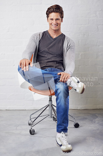 Image of Man, portrait and smile on chair with style on wall background for fashion, confidence or relax. Male person, face and sitting or casual in jeans in studio for happiness in Canada, clothes or outfit