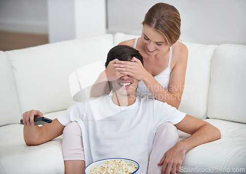 Image of Couple, popcorn and covered eyes with sofa, surprise and remote for playful movie date. People, living room and film for relaxation, smile and bonding on couch with love and wow at home with break