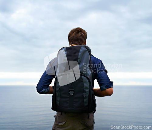 Image of Man, backpack and hiking by ocean for travel, sightseeing or outdoor journey in nature. Rear view of male person, hiker or tourist with bag on back for trekking, fitness or adventure with cloudy sky