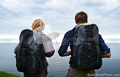 Image of Couple, backpack and hiking by ocean for travel, sightseeing or outdoor journey in nature. Rear view of man, woman or hikers carrying bag on back for trekking, fitness or adventure with cloudy sky