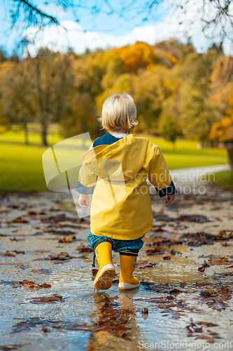 Image of Sun always shines after the rain. Small bond infant boy wearing yellow rubber boots and yellow waterproof raincoat walking in puddles in city park on sunny rainy day.