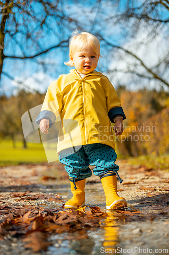 Image of Sun always shines after the rain. Small bond infant boy wearing yellow rubber boots and yellow waterproof raincoat walking in puddles in city park on sunny rainy day.