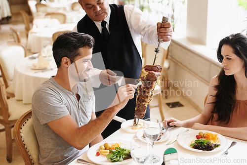 Image of Happy couple, date and waiter with kebab for serving, fine dinning or romantic dinner at table. Young man and woman with chef cutting skewer of food for meal, eating or enjoying service at restaurant