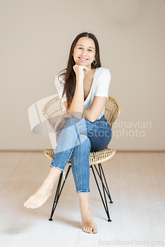 Image of Portrait of confident beautiful woman with long brown hair, wearing casual clothes, sitting on chair in tight jeans and white t-shirt, studio background
