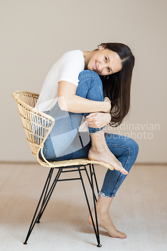 Image of Portrait of confident beautiful woman with long brown hair, wearing casual clothes, sitting on chair in tight jeans and white t-shirt, studio background