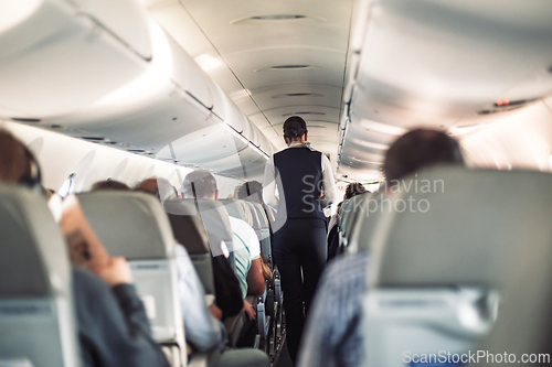 Image of Interior of airplane with passengers on seats and stewardess in uniform walking the aisle, serving people. Commercial economy flight service concept.