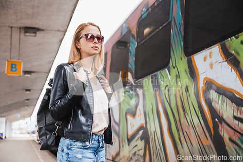 Image of Young blond woman in jeans, shirt and leather jacket wearing bag and sunglass, embarking modern speed train on train station platform. Travel and transportation.