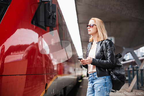 Image of Young blond woman in jeans, shirt and leather jacket wearing bag and sunglass, embarking red modern speed train on train station platform. Travel and transportation.