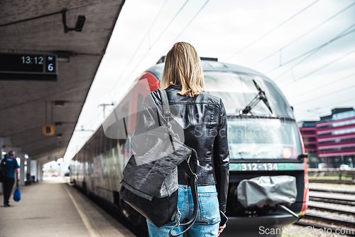 Image of Young blond woman in jeans, shirt and leather jacket wearing bag and sunglass, embarking red modern speed train on train station platform. Travel and transportation.
