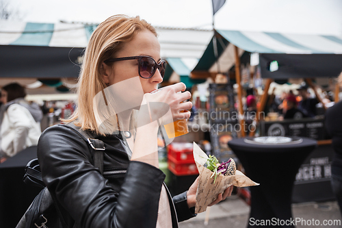 Image of Beautiful young woman holding delicious organic salmon vegetarian burger and drinking homebrewed IPA beer on open air beer an burger urban street food festival in Ljubljana, Slovenia.