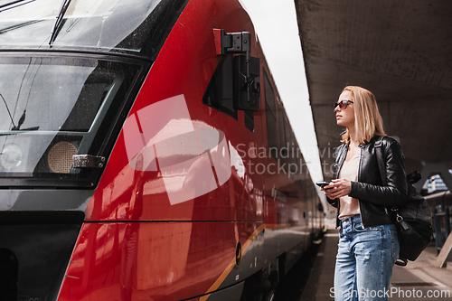 Image of Young blond woman in jeans, shirt and leather jacket wearing bag and sunglass, embarking red modern speed train on train station platform. Travel and transportation.