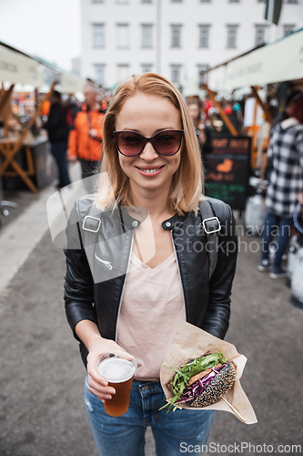 Image of Beautiful young woman holding delicious organic salmon vegetarian burger and homebrewed IPA beer on open air beer an burger urban street food festival in Ljubljana, Slovenia.