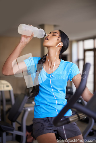 Image of Woman, drinking water and bike in gym, cycling and stationary machine for workout at club. Female person, earphones and hydrate for strength training, music and equipment for fitness and body health