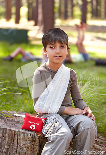 Image of Child, portrait and first aid for hurt arm with sling on bruise for medical treatment outdoors on tree trunk. Kid, emergency kit and bandage for muscle sprain, injury and camp accident in nature.