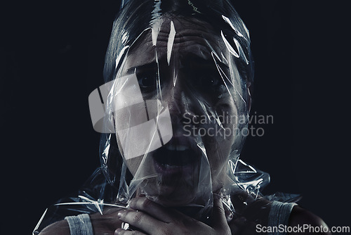Image of Scary, face and woman with plastic or bag in horror, drama or death in dark background of studio. Suffocating, murder and girl with fear from crime, victim and trapped in trash, garbage or pollution