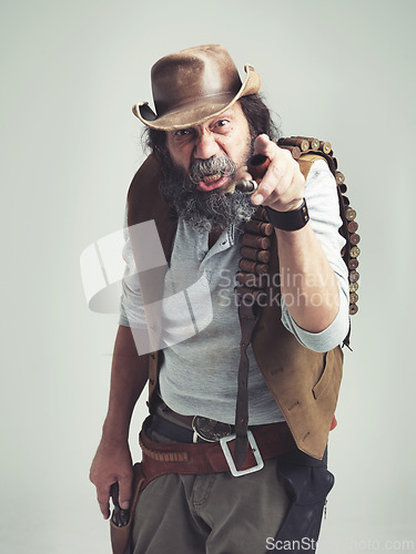 Image of Senior cowboy, point and angry face at studio, costume and holster for western portrait looking scruffy. Old man, sheriff hat and dirty male for texas, grey background and annoyed gesture for outlaw